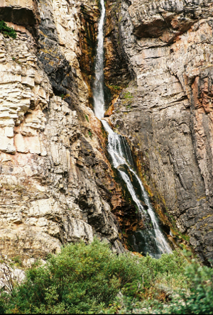 [A thin ribbon of white water runs down a very tall rock face. The rock right around the water area is a darker brown than the rock further out.]